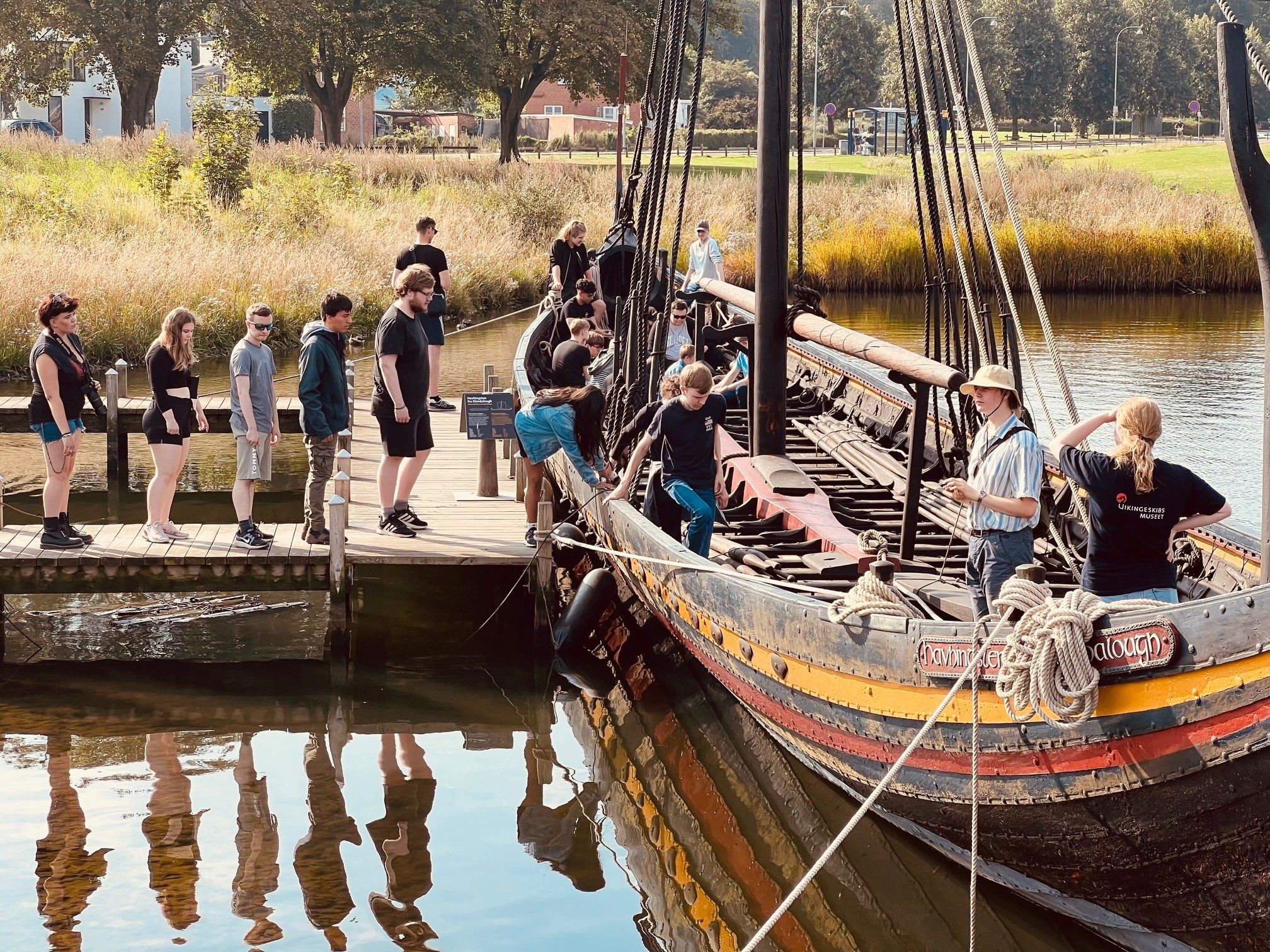 En gruppe unge står på en træbro og går ombord på et vikingeskib ved Roskilde Fjord. Solen skinner, og spejlingen i vandet viser de ventende deltagere.