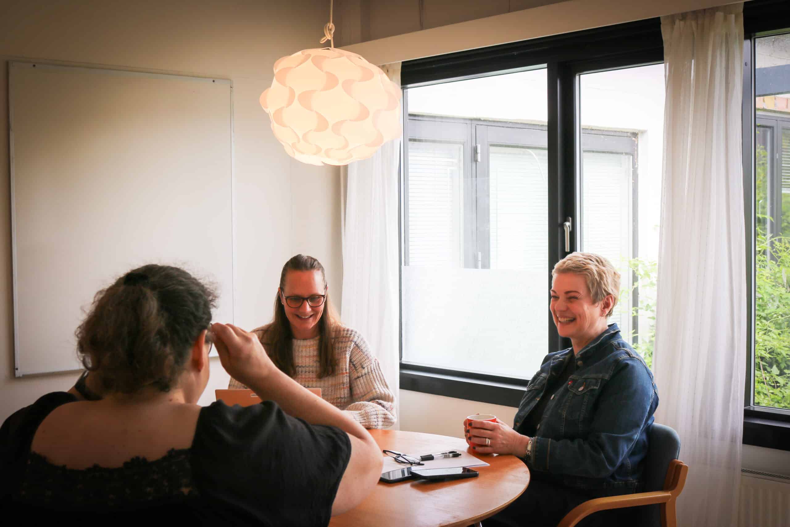 Tre personer sidder omkring et bord i et lyst mødelokale på Center for Autisme. De smiler og taler sammen i en uformel og positiv atmosfære.