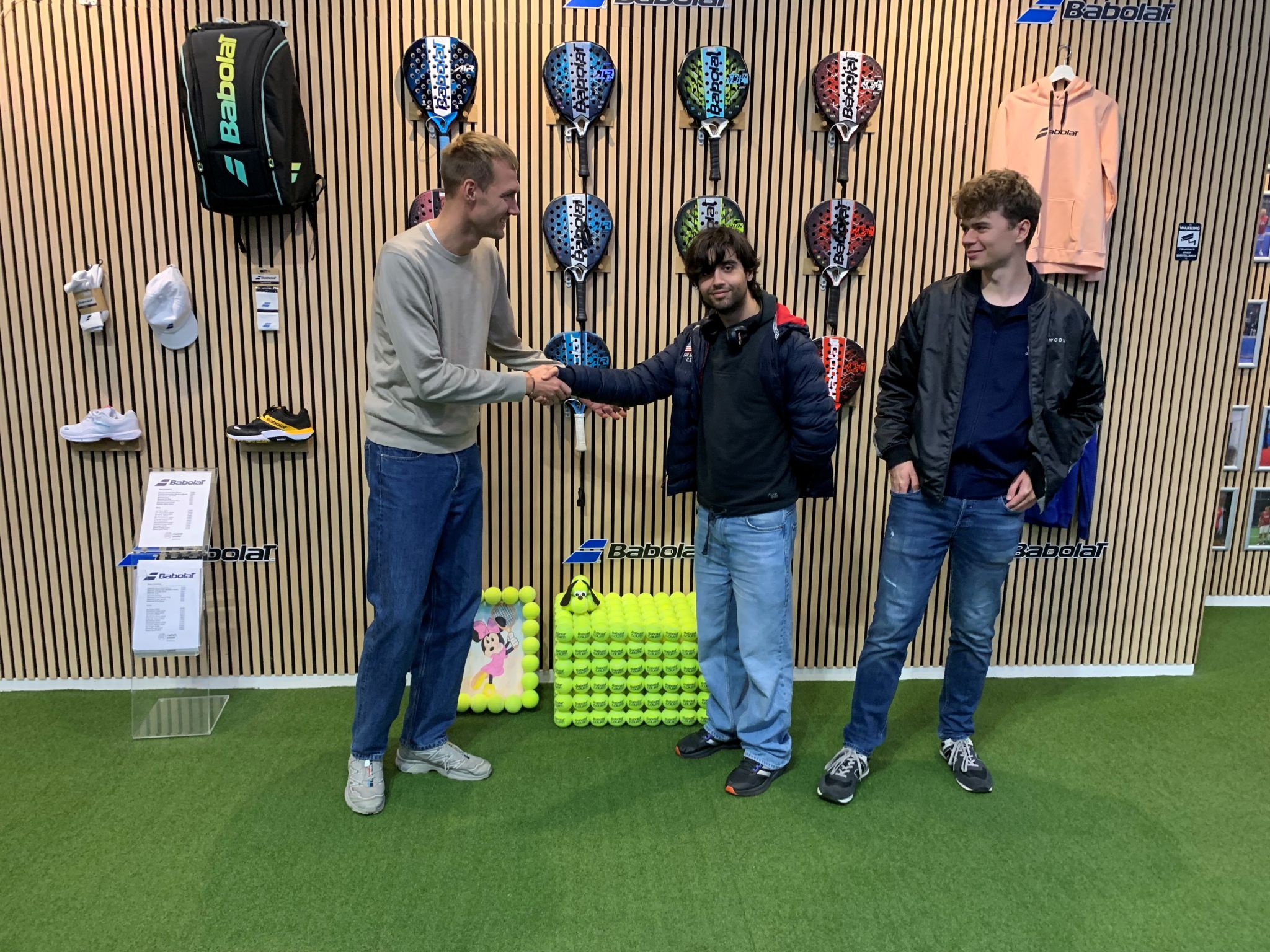Overdragelse af installationskunst fra AVA til Match Padel Danmark i Ballerup. To mænd giver hånd foran en væg med padeludstyr fra Babolat, mens en tredje ser til. Samarbejdet handler om at upcycle brugte padelbolde til kunst skabt af deltagere fra AVA’s kreaværksted for mennesker med autisme.